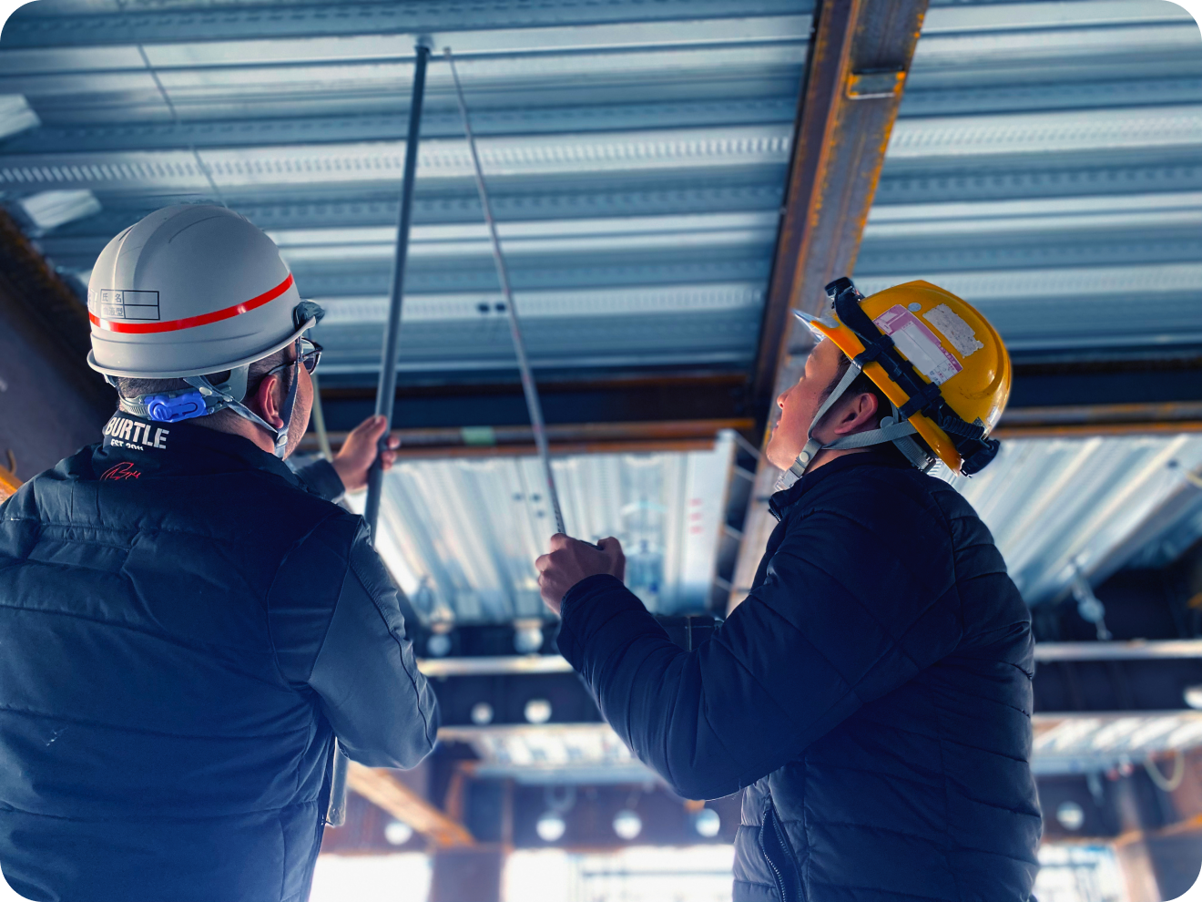 建設現場の天井付近で、協力して吊りボルトの計測作業を行う2人のヘルメット姿の男性作業員
