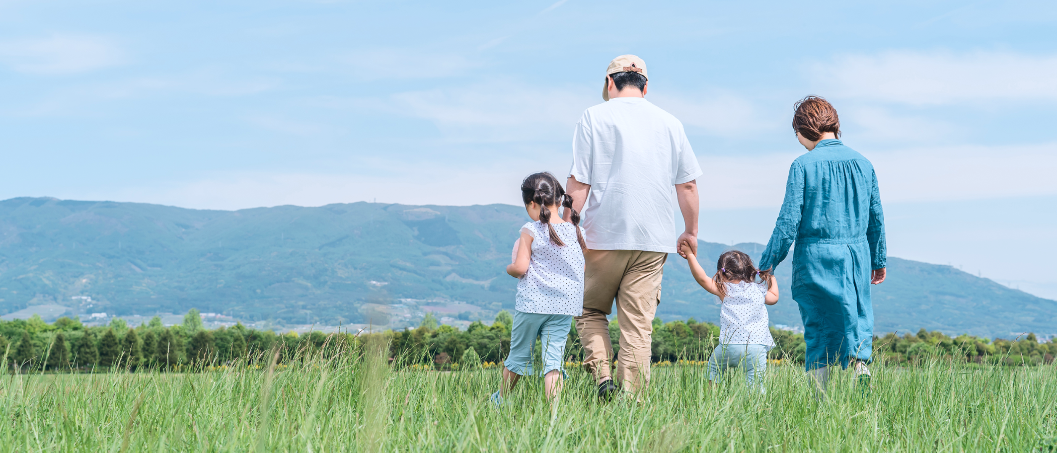 青空と山を背景に、草原を手をつないで歩く両親と二人の子供の後ろ姿