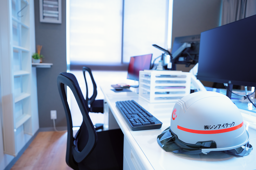 白いデスク上のモニターとキーボードの横に『シンアイテック』の文字が入った安全ヘルメットが置かれている