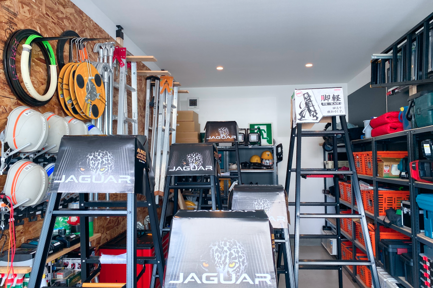 脚立、ケーブル、ヘルメットなどの工具類が棚と壁に整理されて並ぶ倉庫内の様子