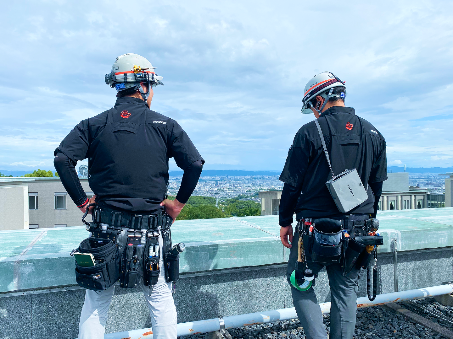 屋上で背を向けて立つ2人の作業員。ヘルメットと黒い作業服を着用し、腰に多数の工具を装備している。手前には建物の縁があり、遠景に市街地と空が広がっている。